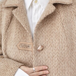 May include: A close-up of a beige wool coat with a single button fastening. The coat has a textured pattern and a soft, fuzzy feel.