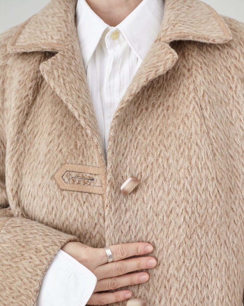 May include: A close-up of a beige wool coat with a single button fastening. The coat has a textured pattern and a soft, fuzzy feel.