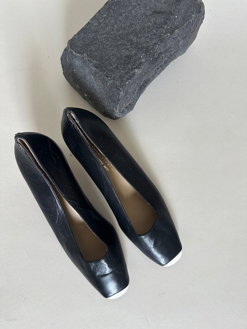 K&ouml;nnte beinhalten: Ein Paar schwarze Lederschuhe mit Absatz, quadratischer Spitze und wei&szlig;em Besatz. Die Schuhe haben eine spitze &Ouml;ffnung an der Oberseite.