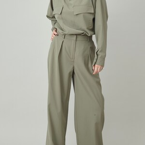 May include: A sage green, long-sleeved collared shirt with a single large pocket on the front. The shirt is tucked into a pair of wide-leg, sage green pants.