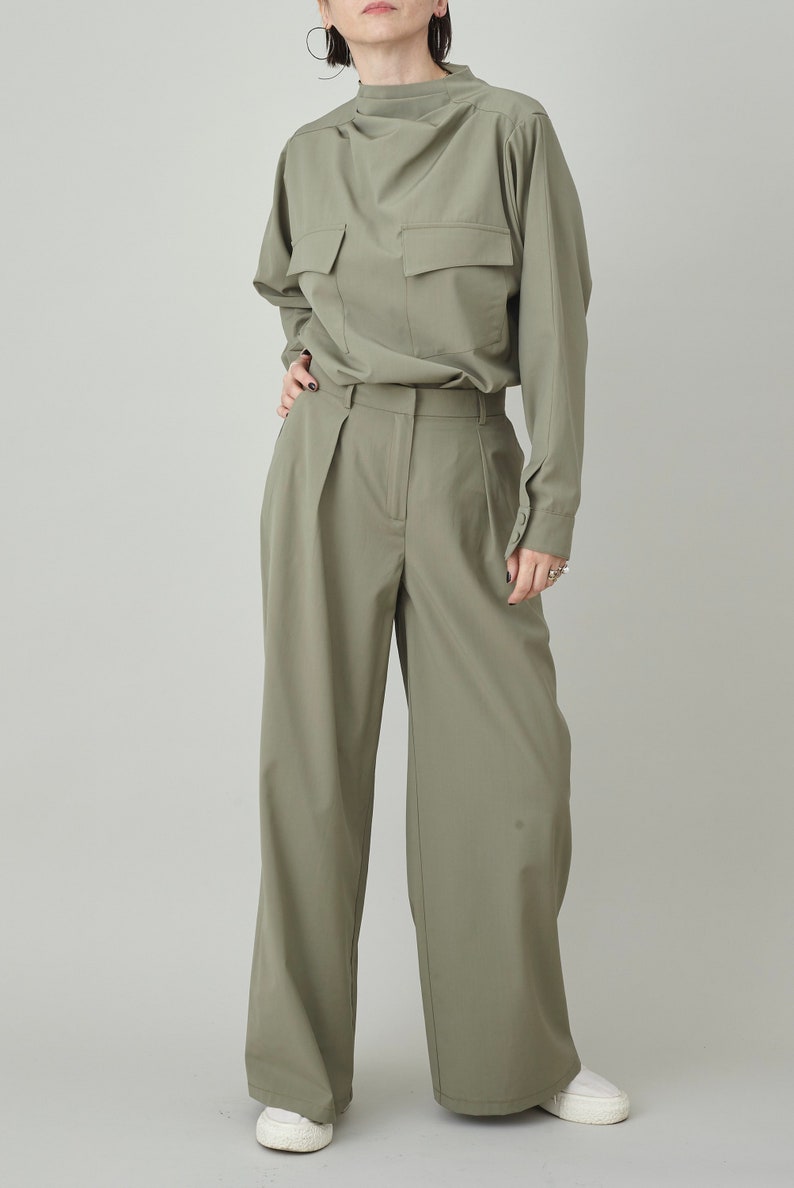 May include: A sage green, long-sleeved collared shirt with a single large pocket on the front. The shirt is tucked into a pair of wide-leg, sage green pants.