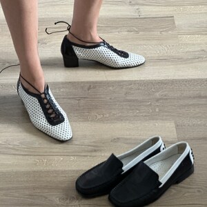May include: Two pairs of shoes. One pair is black leather loafers with white trim. The other pair is white leather shoes with black trim and laces. The shoes are on a wood floor.