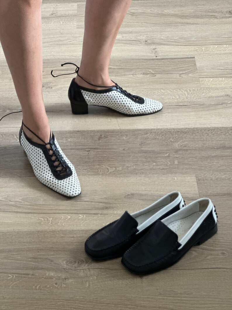 May include: Two pairs of shoes. One pair is black leather loafers with white trim. The other pair is white leather shoes with black trim and laces. The shoes are on a wood floor.