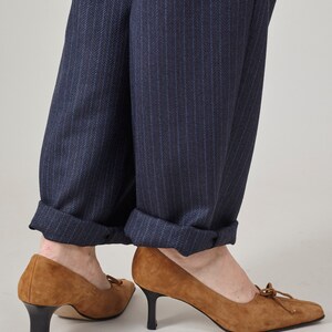 K&ouml;nnte beinhalten: Ein Paar braune Wildlederschuhe mit Absatz und Schleife an der Spitze. Die Schuhe werden zu einer dunkelblauen Hose mit Nadelstreifen getragen.