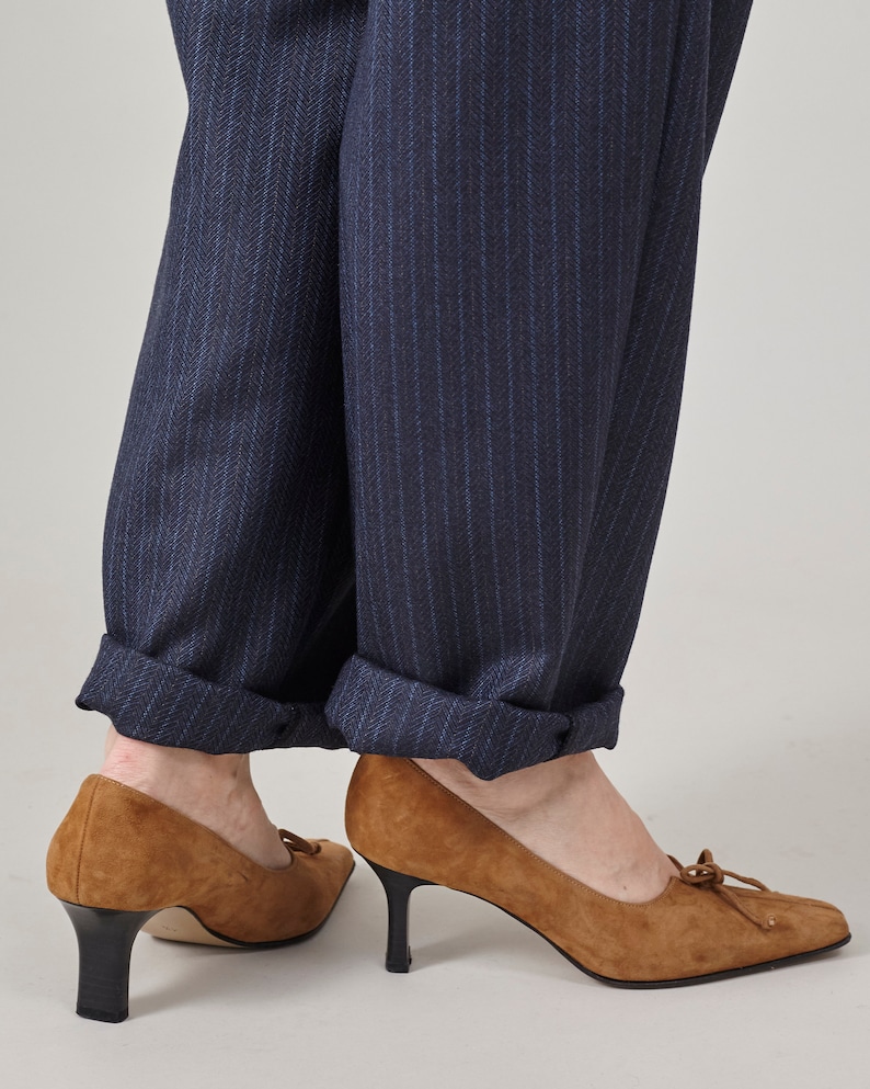 K&ouml;nnte beinhalten: Ein Paar braune Wildlederschuhe mit Absatz und Schleife an der Spitze. Die Schuhe werden zu einer dunkelblauen Hose mit Nadelstreifen getragen.