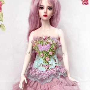 May include: A doll with pink hair and a floral-themed dress. The dress features a light blue bodice with a pink floral pattern and a pink tulle skirt. The bodice is adorned with a pink bow and floral embellishments.