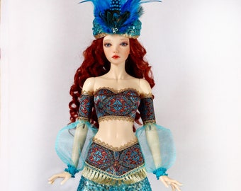 Op de afbeelding: Een pop met een turquoise paillettenrok en een bijpassende top met ingewikkelde gouden en rode patronen. De pop heeft een blauwe gevederde hoofdtooi en lang rood haar. De outfit heeft doorschijnende mouwen en gouden randen.