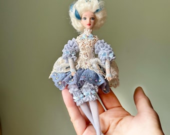 Puede incluir: Una muñeca en miniatura con una peluca blanca con detalles azules y tez pálida. La muñeca viste un vestido azul claro y crema con encaje y mangas con volantes. El atuendo de la muñeca incluye una falda a juego y bombachos con volantes. La muñeca mide unos 15 cm de alto.