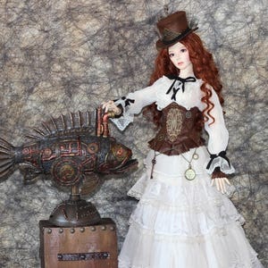 Op de afbeelding: Een steampunk-thema pop met rood haar en een bruine hoge hoed, gekleed in een witte jurk met een bruin korset. Naast de pop staat een decoratief metalen visbeeld met koperkleurige accenten, gemonteerd op een houten voet.