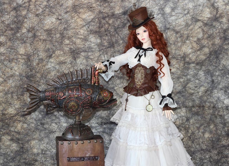 Op de afbeelding: Een steampunk-thema pop met rood haar en een bruine hoge hoed, gekleed in een witte jurk met een bruin korset. Naast de pop staat een decoratief metalen visbeeld met koperkleurige accenten, gemonteerd op een houten voet.