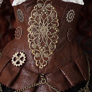 Op de afbeelding: Een bruin steampunk-stijl korset met ingewikkelde gouden filigraan- en tandwielaccenten. Het korset heeft een textuurachtig leerachtig materiaal, een geplooid onderste gedeelte en een delicate ketting. Een witte kanten kraag is zichtbaar.
