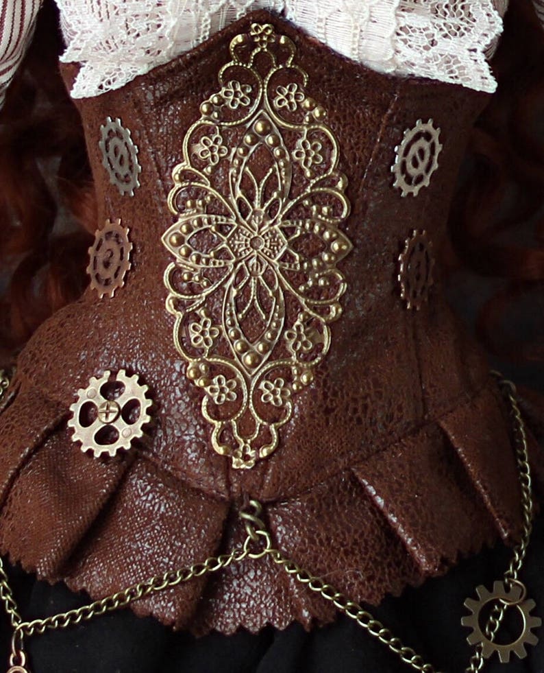 Op de afbeelding: Een bruin steampunk-stijl korset met ingewikkelde gouden filigraan- en tandwielaccenten. Het korset heeft een textuurachtig leerachtig materiaal, een geplooid onderste gedeelte en een delicate ketting. Een witte kanten kraag is zichtbaar.