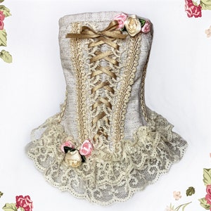 May include: A decorative corset-shaped item in a neutral color, featuring gold-colored lace trim and a matching ribbon bow. The design includes a lace-up front with gold ribbon and is adorned with small pink and cream-colored roses. The bottom edge has a delicate lace trim.