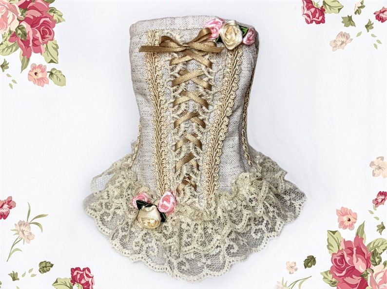 May include: A decorative corset-shaped item in a neutral color, featuring gold-colored lace trim and a matching ribbon bow. The design includes a lace-up front with gold ribbon and is adorned with small pink and cream-colored roses. The bottom edge has a delicate lace trim.