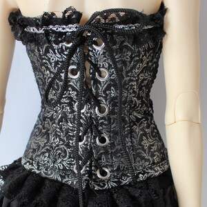 May include: Black and silver corset with a floral pattern, lace trim, and a laced front. The corset has silver eyelets and a black cord. The garment is part of a gothic or Victorian-inspired ensemble.