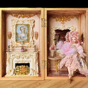 May include: A miniature room box featuring a fireplace with a gold screen, a framed portrait, and a doll with pink hair wearing a ruffled dress. The scene includes a small table, a mirror, and gold decorative accents within a wooden structure.