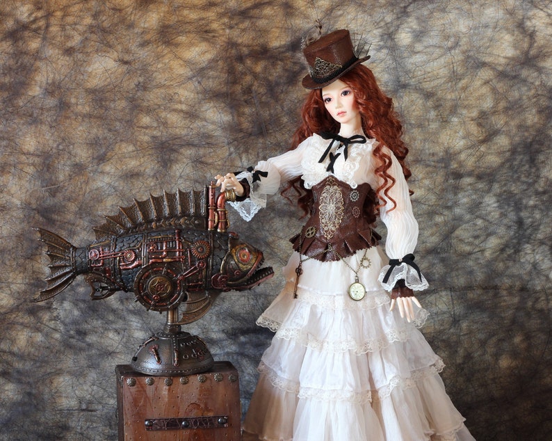 Op de afbeelding: Een steampunk-thema pop met lang rood haar, gekleed in een witte jurk, bruin korset en een kleine hoge hoed. De pop staat naast een gedetailleerde, bronskleurige vissculptuur met mechanische elementen, gemonteerd op een houten standaard.