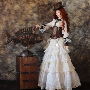 Op de afbeelding: Een steampunk-pop met rood haar, een bruine hoge hoed, een witte ruchesjurk en een bruin korset. Naast de pop staat een gedetailleerde, mechanische vissculptuur op een houten kist, die Victoriaanse en industri&euml;le elementen combineert.
