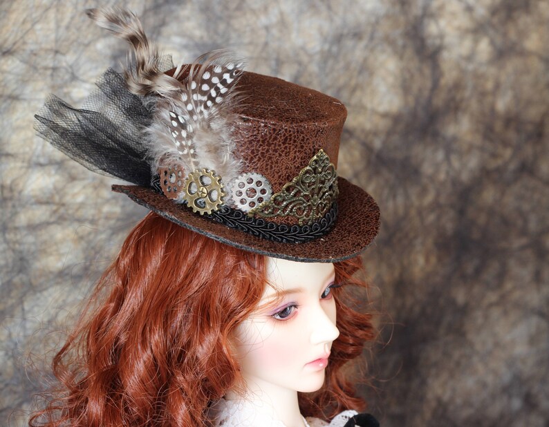 Op de afbeelding: Een bruine steampunk-stijl hoge hoed met een getextureerde afwerking. De hoed is versierd met veren, zwart gaas en decoratieve tandwielen. Een gouden filigraanontwerp accentueert de voorkant. Een uniek accessoire voor cosplay.
