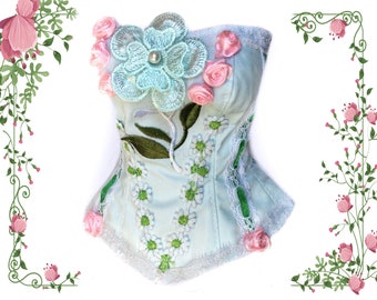 May include: A light blue corset with floral embroidery. The corset features a large, embroidered turquoise flower with a pearl center, pink roses, and white daisy-like flowers. Green leaves and trim add detail to the design.