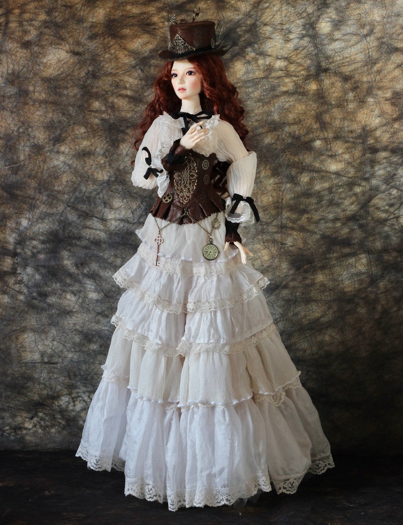 Op de afbeelding: Een pop gekleed in een steampunk-ge&iuml;nspireerde outfit. De pop heeft lang rood haar en draagt een bruine hoge hoed, een witte blouse met zwarte accenten en een bruin korset. De rok is een gelaagde, gerimpelde witte stof met kanten rand.