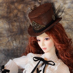Op de afbeelding: Een bruine steampunk-stijl hoge hoed met decoratieve versieringen, veren en zwart gaas. De hoed wordt gedragen door een pop met lang, rood haar en een witte kanten blouse met een zwarte strik.