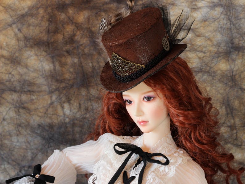 Op de afbeelding: Een bruine steampunk-stijl hoge hoed met decoratieve versieringen, veren en zwart gaas. De hoed wordt gedragen door een pop met lang, rood haar en een witte kanten blouse met een zwarte strik.
