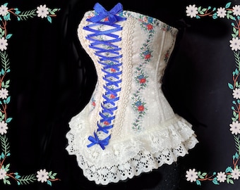 May include: A cream-colored corset with a floral pattern of red and blue flowers. The corset features blue ribbon lacing and bows, with a lace trim at the bottom. The background is black.