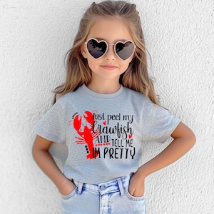 Puede incluir: Camiseta gris claro con un diseño de cangrejo rojo y el texto "Just peel my crawfish and tell me I'm pretty". La persona lleva gafas de sol en forma de corazón.
