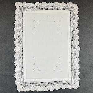 May include: White rectangular tablecloth with a decorative crocheted lace border. The centre features a cutwork design with floral and dot patterns. The tablecloth is made of a soft, white fabric, suitable for a dining table or decorative display.