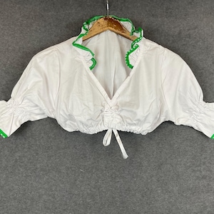 May include: White cropped blouse with green trim and ruffled neckline. The blouse has a V-neck and gathered details at the waist and sleeves. It is hanging on a wooden hanger.