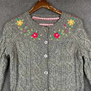 May include: A grey cable-knit cardigan featuring a round neckline with embroidered yellow, red, and green floral patterns. The garment includes a front button closure with five buttons and a red and white checkered fabric trim along the inner collar.