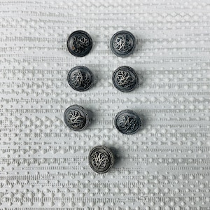 Peut inclure: Sept boutons métalliques circulaires disposés en motif vertical. Chaque bouton présente une finition argentée oxydée foncée avec un motif de monogramme complexe en relief au centre, entouré d'une bordure perlée texturée sur le bord extérieur.