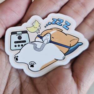 Puede incluir: Una pegatina con un corgi de dibujos animados durmiendo con una máquina CPAP y un pájaro. El corgi está sobre una almohada azul con "zzz" sobre su cabeza. La máquina CPAP está etiquetada como "APNEA".