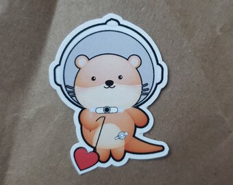 May include: A cartoon otter wearing a space helmet and holding a red heart. The otter is wearing a white space suit with a black belt and a black circle on the chest. The otter is standing on a brown background.
