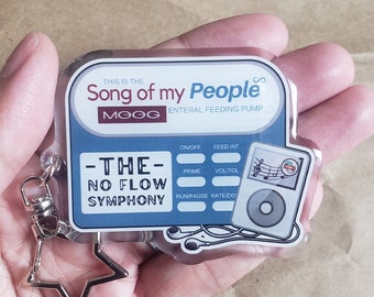 May include: A blue and white keychain with a star charm. The keychain features a design of a portable music player with the text "This is the Song of my People MOOG Enteral Feeding Pump -THE- NO FLOW SYMPHONY".