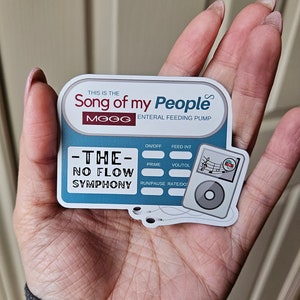 May include: A blue and white sticker with the text "This is the Song of my People" and "MOOG Enteral Feeding Pump". The sticker also has a graphic of a medical pump with the text "The No Flow Symphony" and various buttons labeled "On/Off", "Feed Int", "Prime", "Vol/Tol", "Run/Pause", and "Rate/Do".