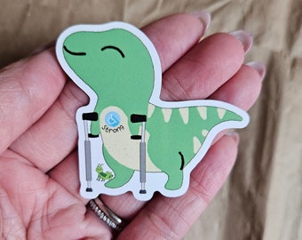 Pode incluir: Adesivo de dinossauro verde com um rosto sorridente, segurando muletas e usando um símbolo de cadeira de rodas azul e branco com a palavra "strong" escrita abaixo. Também é possível ver um pequeno inseto verde no adesivo.