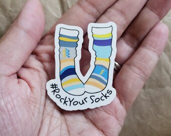 May include: A sticker depicting two socks with a colourful design of stripes and geometric shapes in blue, yellow, and grey. The text "#RockYourSocks" is included. The sticker is held in a person's hand.