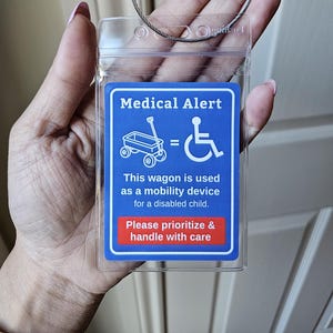 May include: A clear plastic medical alert tag with a blue and red label. The label features a wagon and wheelchair symbol, with text stating the wagon is a mobility device for a disabled child. Includes a metal loop for attachment.