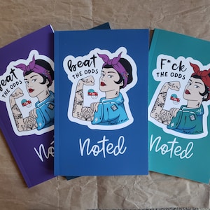 May include: Three notebooks in purple, blue, and teal. Each notebook features a sticker of a woman flexing her arm with the text "beat the odds" or "f*ck the odds". The word "Noted" is written in cursive below the sticker.