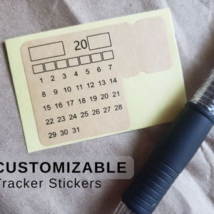 Peut inclure: Un autocollant rectangulaire beige présentant une grille de calendrier mensuel imprimée avec les chiffres de 1 à 31, un espace pour l'année 20 et des cases vides pour des notes. L'autocollant est placé à côté d'un stylo noir à grip texturé.