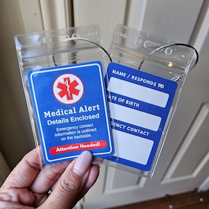 Puede incluir: Un portagafete de plástico transparente con una tarjeta azul en su interior. La tarjeta tiene un símbolo rojo de estrella de la vida y el texto "Medical Alert Details Enclosed". La tarjeta también tiene el texto "Emergency contact information is outlined on the backside. Attention Needed!"