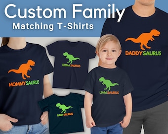 Op de afbeelding: Een collectie marineblauwe t-shirts met dinosaurusafbeeldingen en tekst. De shirts tonen de teksten MOMMYSAURUS, DADDYSAURUS, EMMA SAURUS, LIAM SAURUS en BABYSAURUS in oranje en groen, met bijbehorende oranje of groene silhouetten van dinosaurussen.