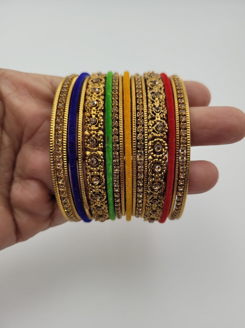 Puede incluir: Un conjunto de pulseras doradas con intrincados dise&ntilde;os florales y gemas incrustadas. El conjunto incluye pulseras en varios colores, como azul, verde, amarillo y rojo, creando un aspecto vibrante y ornamentado. Las pulseras se sostienen en una mano.