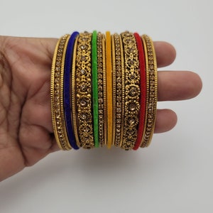 Puede incluir: Un conjunto de pulseras doradas con intrincados dise&ntilde;os florales y pedrer&iacute;a incrustada. Las pulseras est&aacute;n acentuadas con bandas de tela azul, verde, amarilla y roja. Las joyas se exhiben sobre un fondo blanco.