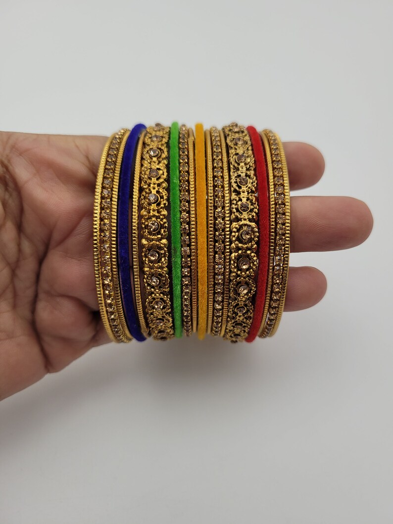Puede incluir: Un conjunto de pulseras doradas con intrincados dise&ntilde;os florales y pedrer&iacute;a incrustada. Las pulseras est&aacute;n acentuadas con bandas de tela azul, verde, amarilla y roja. Las joyas se exhiben sobre un fondo blanco.
