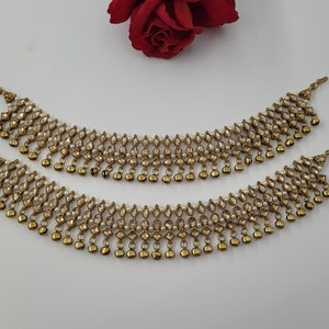 Op de afbeelding: Twee goudkleurige enkelbandjes met een herhalend patroon van heldere en amberkleurige edelstenen en kleine, bungelende gouden belletjes. De enkelbandjes worden gepresenteerd op een wit oppervlak met een rode roos op de achtergrond. Sieraden.