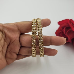 May include: Two gold-colored bangles with oval-shaped, light-colored stones. The bangles are held in a hand against a white background, with a red rose in the background. The jewelry is likely for women.