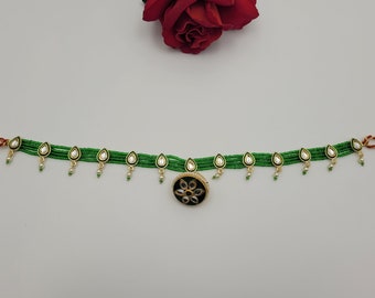 May include: A green beaded belt with gold detailing and teardrop-shaped pendants. The belt has a central black and gold floral design. Small white pearls hang from the teardrop pendants. The belt is shown against a white backdrop.